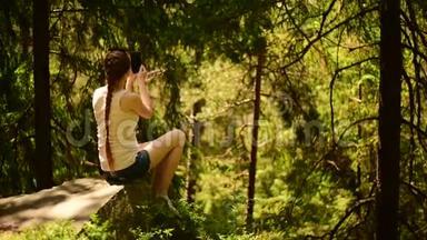美丽的女孩坐在石头上，自拍，在智能手机上拍摄视频。 针叶林，晴天